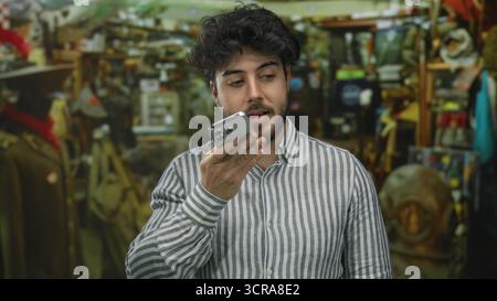 Young hispanic man with beard recording voice message on smartphone in indoor bazar surrounded by eclectic items and wearing striped shirt. Stock Photo