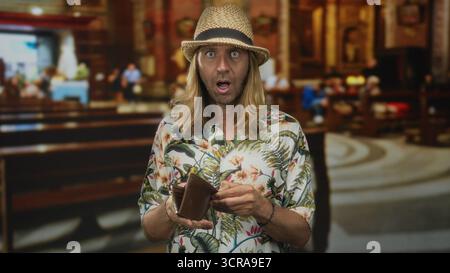 Blond man wearing straw hat holds open wallet in church building with surprised expression; surprise. Stock Photo
