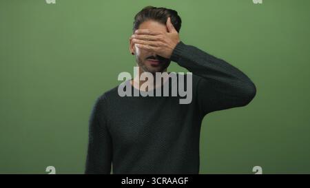 Young bearded man covers eyes with hand while smiling in studio; shyness mystery privacy playfulness. Stock Photo