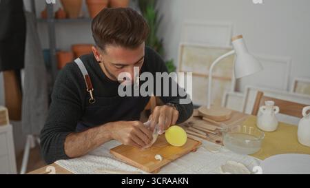 Man shaping clay with his hands in a ceramics studio, kneading soft clay pieces on a wooden board with sculpting tools visible; concentration. Stock Photo