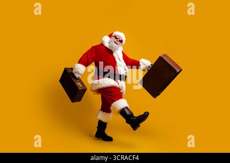 Joyful Santa Claus in vibrant red outfit stylishly carrying travel bags enjoying holiday in front of a bright yellow background Stock Photo