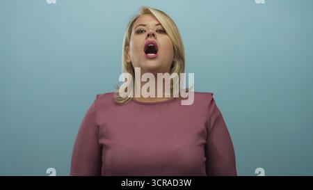 Blonde woman pinches nose wearing a mauve top in a light blue studio set reacting to a foul smell; disgust. Stock Photo