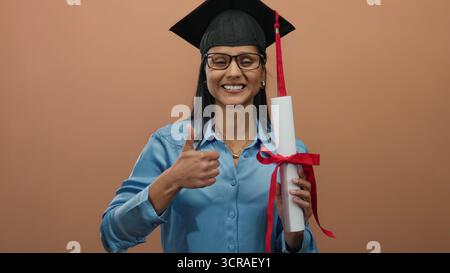 Smiling woman with glasses and graduation cap holding diploma giving thumbs-up in front of brown wall Stock Photo