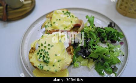 Eggs benedict with creamy hollandaise sauce and fresh salad served on a white plate in a cafeteria setting showcasing a delicious breakfast meal. Stock Photo