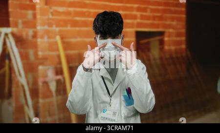 Young man in mask and lab coat at construction site points at camera, indicating attention or focus, blending professional attire with industrial sett Stock Photo