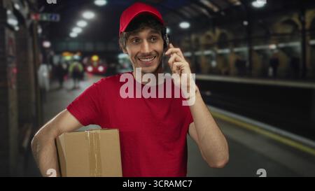 Man holds cardboard box with one arm and talks on phone at train station; efficiency reliability service. Stock Photo