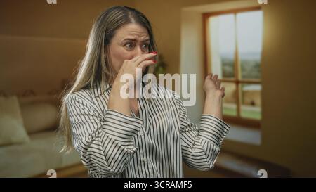 Middle aged blonde woman pinches nose beside window in spacious yellow building lobby wearing striped shirt; disgust. Stock Photo