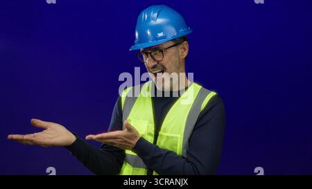 Man in hardhat and safety vest gesturing confidently against a blue background, depicting skilled professional engaging in a presentation or instructi Stock Photo