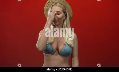 Woman with blonde hair wearing straw hat and blue bikini top pinches nose on red studio set; disgust. Stock Photo