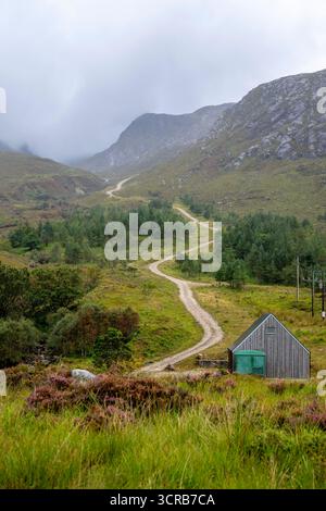 A tranquil landscape of rolling hills, a dirt path winds through a pine forest toward a rustic wooden hut, with rugged mountains and low clouds creati Stock Photo