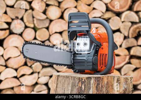 Closeup portable gasoline chainsaw on wood chunk in mosaic background of logs. Modern sawing machine with replaceable chain before blur firewood wall. Stock Photo