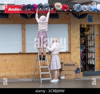 Eine Standlfrau mit ihrer Kollegin bringt ihre Ware Hüte von außen mit einer Stehleiter an der Andenken und Souvenir Bude an am Morgen vor Eröffnung des Oktoberfestes bei verregnetem Wetter Wolken Regen Schlechtwetter auf der Wiesn Oktoberfest in München / Datum: 30.09.2025 / *** A stand woman with her colleague brings her goods hats from the outside with a stepladder to the souvenir stall on the morning before the opening of the Oktoberfest in rainy weather Clouds Rain Bad weather at the Oktoberfest Oktoberfest in Munich Date 30 09 2025 Stock Photo