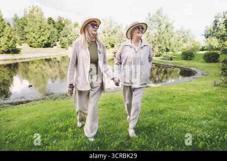 Senior women walking outdoors in stylish beige linen outfits, casual jackets, wide leg trousers, hats and sunglasses, full length fashion portrait sho Stock Photo