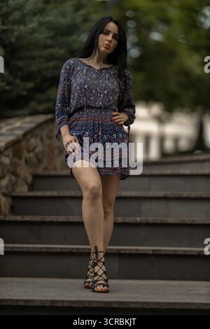 young woman confidently walks down a set of stone steps. She wears a colorful dress and sandals surrounded by greenery in a peaceful garden setting. Stock Photo