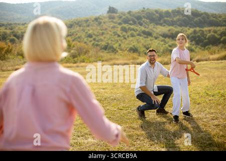 A father and daughter are playing catch in a green field while a woman in a pink shirt runs towards them. The landscape features hills and trees under Stock Photo