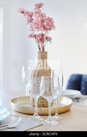 Stylish dining table with clean glass wine glasses, golden tray, and woven vase with pink artificial flowers. Background of interior details with soft Stock Photo