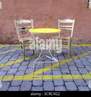 Rustic café set with two weathered chairs and small yellow round table on cobblestone street. Stock Photo