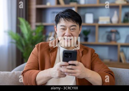 Asian man smiling on a modern living room sofa, relaxed and engaged with his smartphone, browsing social media and messaging for entertainment and connection at home Stock Photo