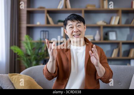 Asian man smiling and gesturing with hands while having an online video call from his home living room, connecting with friends, family, or colleagues Stock Photo