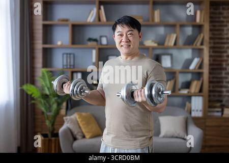 Asian man smiling while doing bicep curls with dumbbells in his living room, focusing on strength training and maintaining a healthy lifestyle through home workouts Stock Photo
