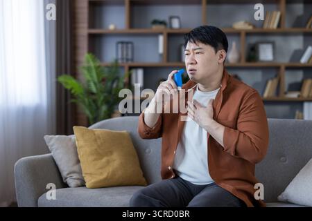 Asian man sitting on a sofa at home using a blue inhaler, experiencing shortness of breath and difficulty breathing during an asthma attack, holding a hand to his chest Stock Photo