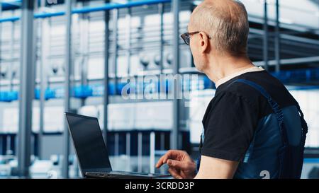 Smart factory expert using laptop software to test automated systems and ensure reliability. Worker uses program on notebook to identify bottlenecks and optimize gear based on sensors data, camera A Stock Photo