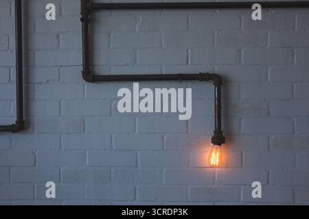 A view of a dimly lit brick wall facade, featuring rusty angled pipes and a glowing vintage light bulb, as a background. Stock Photo