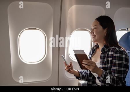 Young Asian woman sitting by the airplane window holding a passport and smartphone, smiling while checking travel documents and enjoying her flight ex Stock Photo