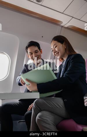 Asian business professionals working together on a laptop while sitting on a commercial airplane. They appear to be discussing a project or presentati Stock Photo