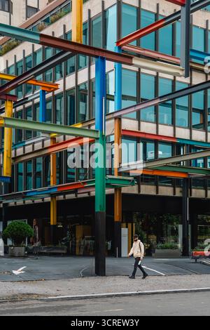 Colorful steel beams on modern glass facade of commercial building in urban street scene, person walking past Stock Photo