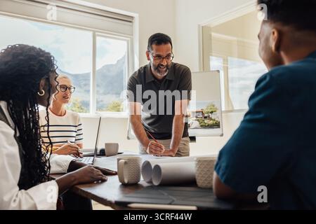 A team of architects discussing project details in a bright and modern workspace. The group includes various ages and genders, fostering creativity an Stock Photo