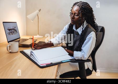 A young Black woman working on tasks at a desk, with a laptop and materials for architecture design. The setting conveys a professional yet creative o Stock Photo