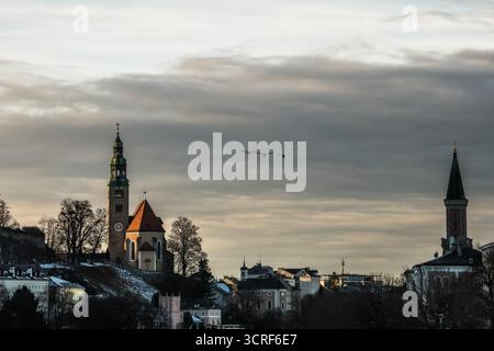 View of the old town's church with a tall green spire and a building with a dark spire, under a cloudy sky with flying birds, Salzburg, Austria. Stock Photo