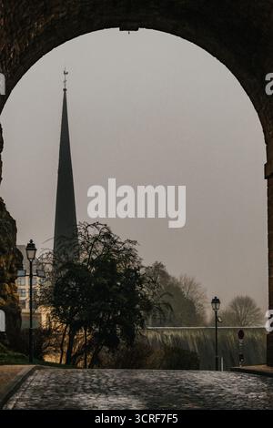 View of a sharp spire piercing the misty sky, framed by the stone archway, with a cobblestone street leading to the scene, Luxembourg city, Luxembourg. Stock Photo