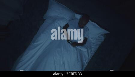 Man in Bed Reads News on Smartphone, While Explosions Outside Window Shake Bedroom. At Night, Man Hides in Panic From Explosions. Air Raid Sirens. Stress, Deprivation Of Sleep. Rotating Top View. Stock Photo