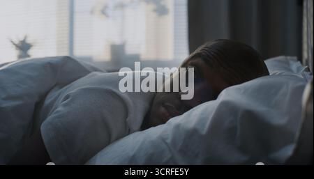 Close up of Adult Guy Slowly Wakes Up in Bed, Resting Head on Soft Pillow, Smiling in Bright Sunlight. Concept of Healthy Rest, Sleep, and Recovery, Healthy Lifestyle. Static Shot. Portrait. Stock Photo