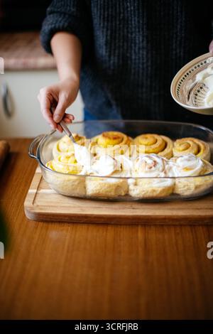 Person spreading smooth white icing on warm cinnamon rolls in a glass dish, fresh from the oven Stock Photo