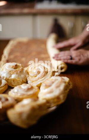 Rolling cinnamon dough on a wooden table with baked rolls in the foreground, capturing the home baking process Stock Photo