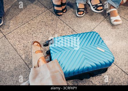 A blue suitcase rests on the airport floor. Nearby, a group of people in casual attire waits. Their feet are visible, showcasing various sandals and s Stock Photo