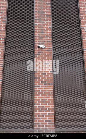 Surveillance camera mounted on a red brick wall with black metal grid panels, symbolizing security, monitoring, and urban architecture. Stock Photo