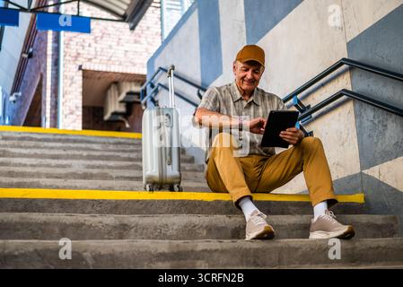 Happy senior man is sitting at railway station and waiting for boarding. He is checking the time on his wristwatch. Senior people traveling. Stock Photo