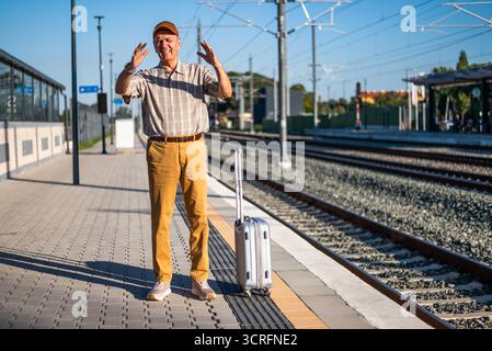 Angry senior man is standing at railway station. He is angry because train is late. Senior people traveling. Stock Photo