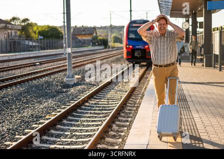 Senior man is standing at railway station. He missed the train departure and holding his head in disbelief. Senior people traveling. Stock Photo