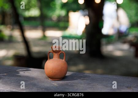 This image showcases a small terracotta vase with two handles, resting on a wooden table amidst a serene outdoor setting rich in greenery and soft lig Stock Photo