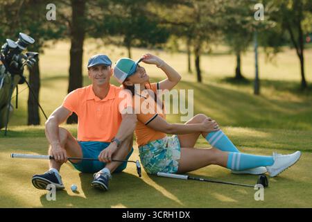 Two golfers relax on the green under the sun. They sit on the grass, enjoying a break while chatting and laughing, with golf clubs and bags nearby. Stock Photo