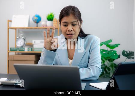 Professional Asian businesswoman gives OK hand gesture during virtual meeting, showing agreement and satisfaction. Symbol of clear communication, team Stock Photo