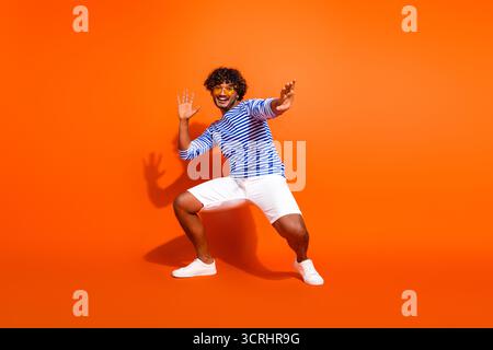 Young stylish man making fun poses in a striped shirt against bright orange background, embodying joy and energy Stock Photo
