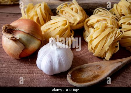 A close up of dried Italian Tagliatelle pasta on the kitchen work top with  onion, garlic and a wooden spoon. Stock Photo