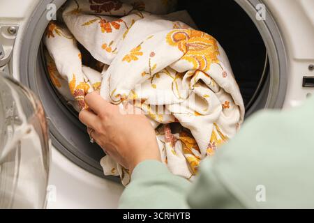 Woman loading beige decorative linen fabric into front load washing machine. Natural textile laundry concept. Home cleaning and fabric care background Stock Photo