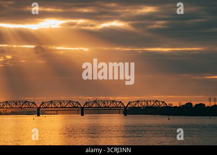 Sunbeams piercing through dramatic clouds over a river, illuminating a silhouetted truss bridge at golden hour, reflecting vibrant orange and yellow hues on the tranquil water surface Stock Photo